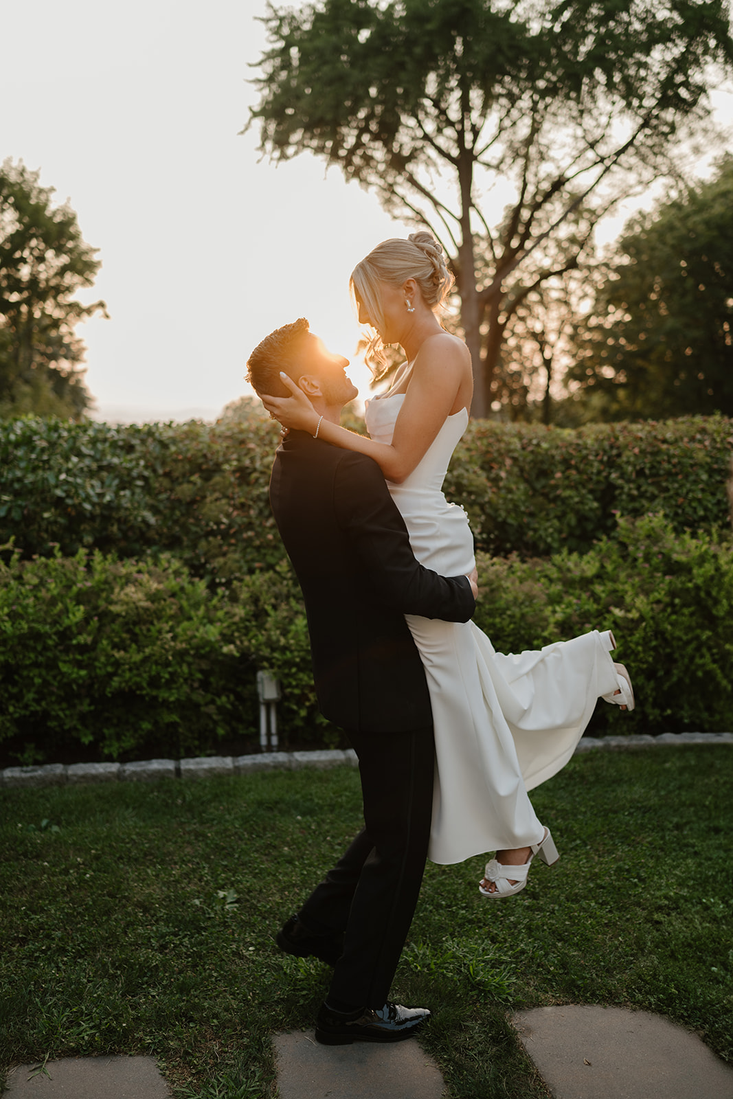 Groom lifts the bride in his arms during a golden sunset, framing them in warm light on the lawn.